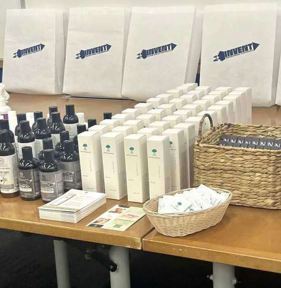 Dr. Ishii — U.S. activation table at the INVENT / GWWIB Spring Conference, featuring Basic Lotion boxes, sample sachets, and partner products